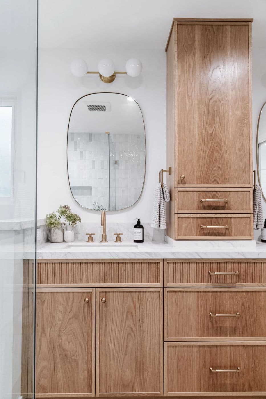 Walnut bathroom vanity and tall cabinet with a round mirror and brass sconces