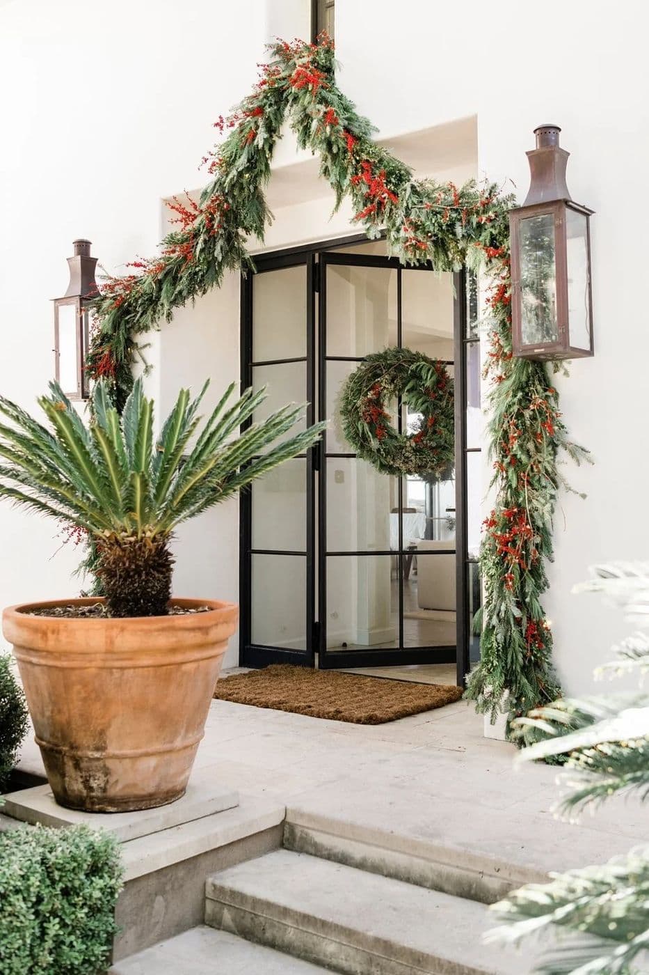 Front door framed in garland with a wreath and potted palm