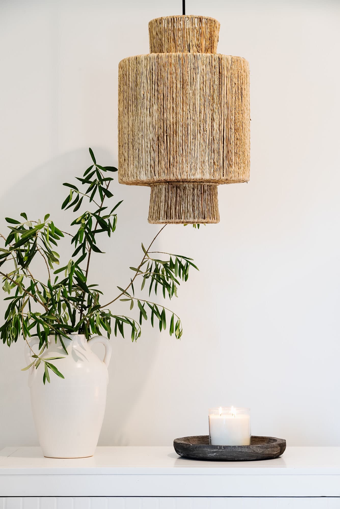 Woven pendant light above a white console styled with an olive branch and a candle