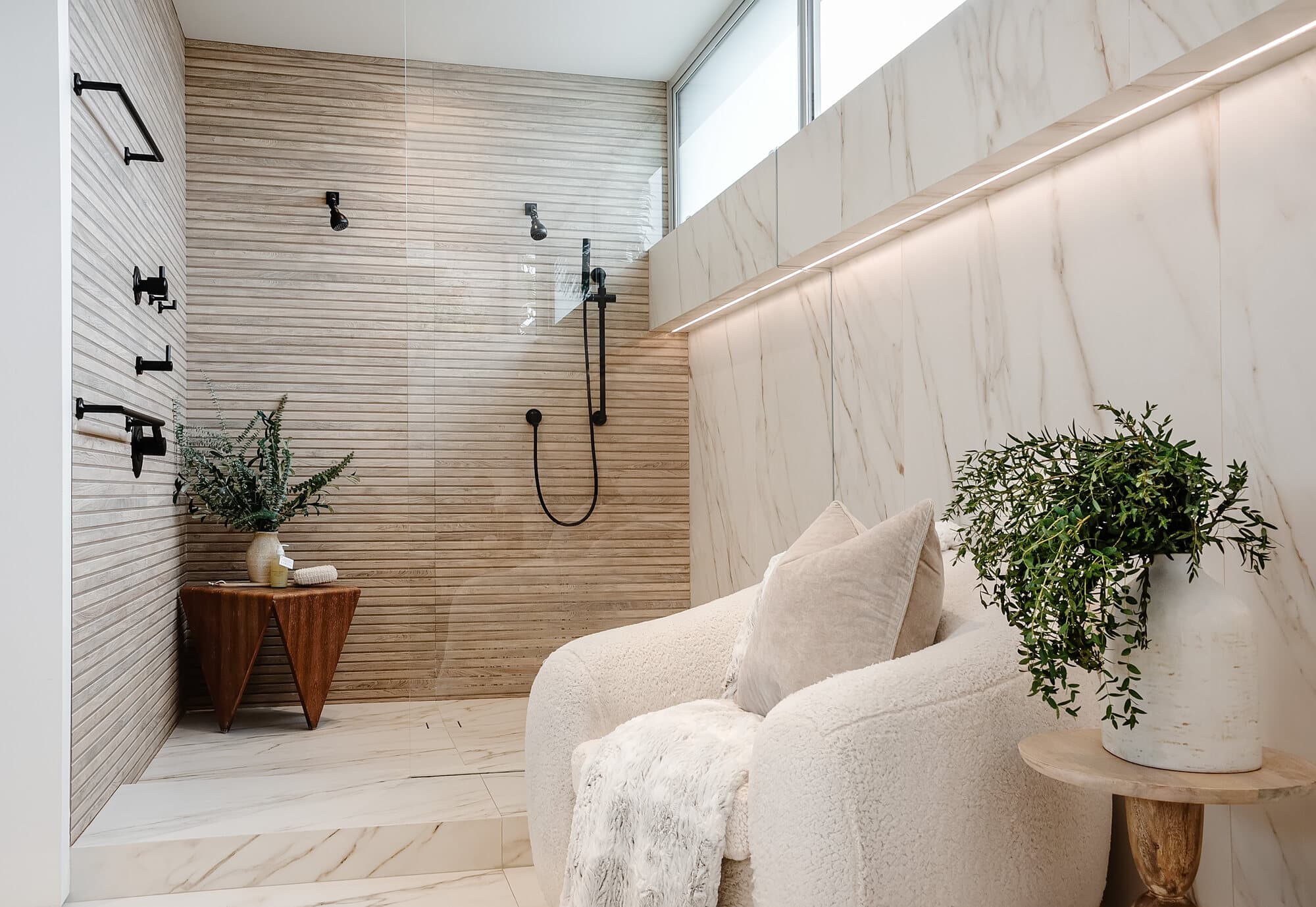 Bright marble walk-in shower with black fixtures beside a boucle chair and greenery