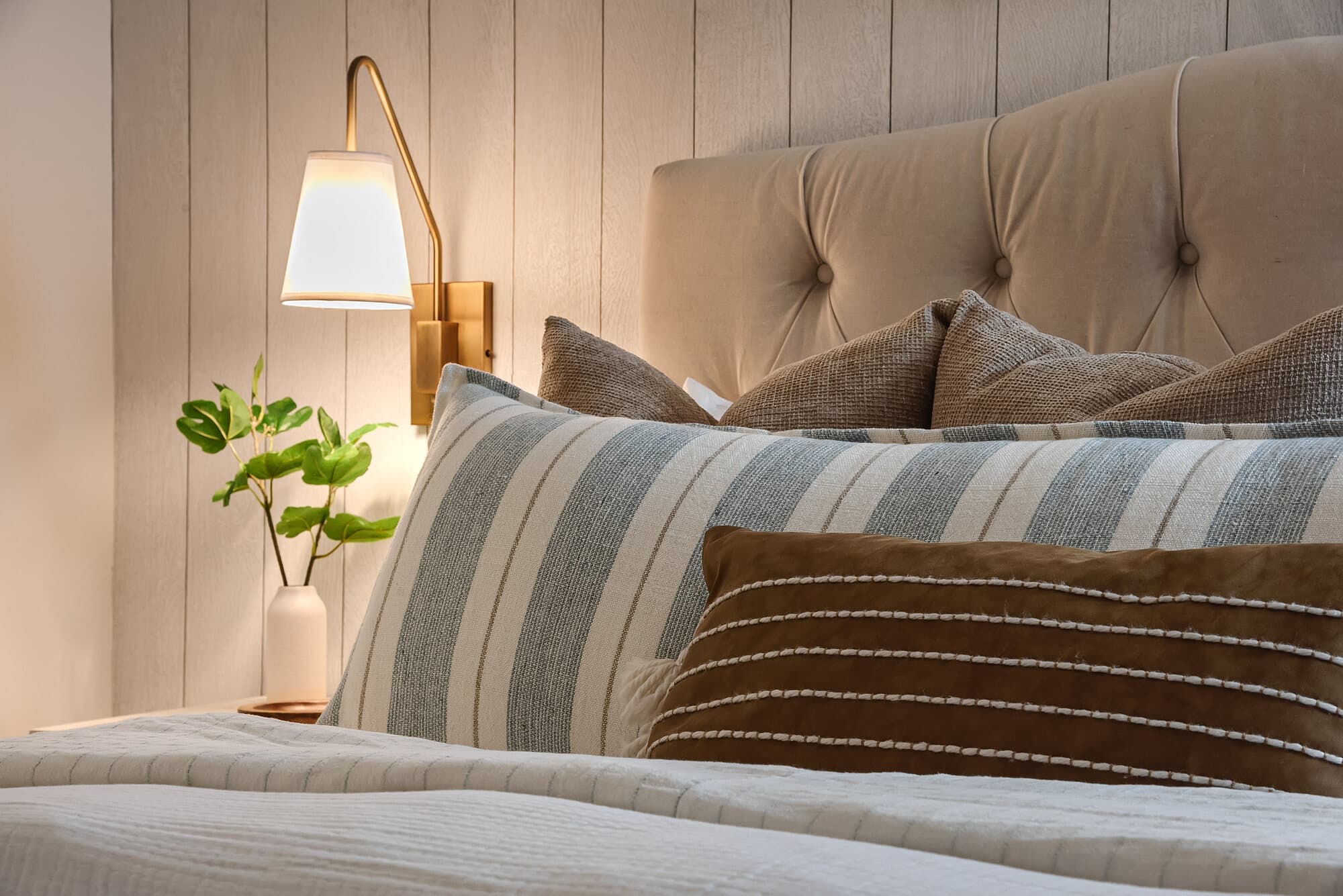 Styled bed with a tufted headboard, layered neutral pillows, and a brass sconce